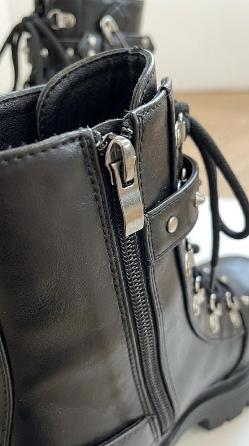 May include: Close-up of a black leather boot with a silver zipper and metal accents. The boot features a lace-up design and a chunky sole, showcasing a modern, edgy style.