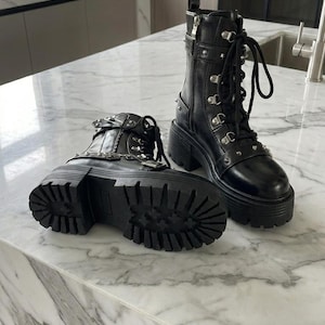 May include: A pair of black leather platform boots with silver-tone hardware, including studs, buckles, and zippers. The boots have thick, lug soles and are laced up the front. The boots are displayed on a marble countertop.