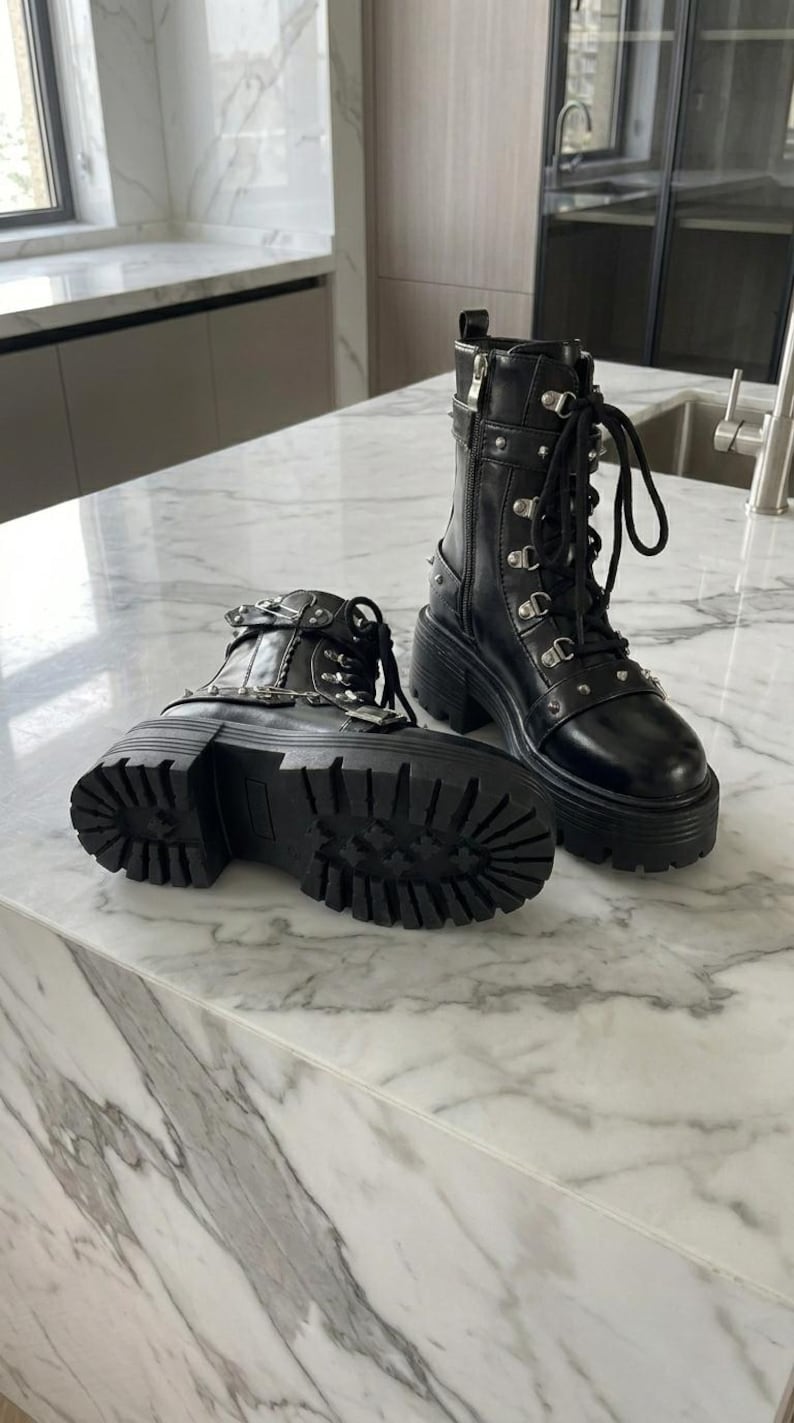 May include: A pair of black leather platform boots with silver-tone hardware, including studs, buckles, and zippers. The boots have thick, lug soles and are laced up the front. The boots are displayed on a marble countertop.