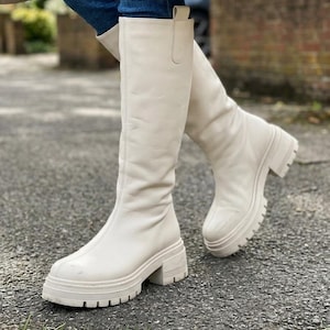 May include: A pair of off-white knee-high boots featuring a smooth leather construction, a rounded toe, a vertical seam detail along the front, and a thick, chunky lug sole with a block heel. The boots include a small pull tab at the top.