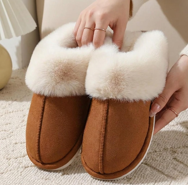 Peut inclure: Une paire de pantoufles en daim marron avec une bordure en fausse fourrure cr&egrave;me. Les pantoufles ont un bout arrondi et une semelle blanche. L'image met en &eacute;vidence la texture douce et le design confortable des pantoufles.