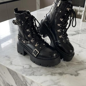 May include: A pair of black leather ankle boots with chunky soles. The boots feature multiple silver-tone metal accents, including buckles, rivets, and eyelets for the laces. The boots are displayed on a marble surface.