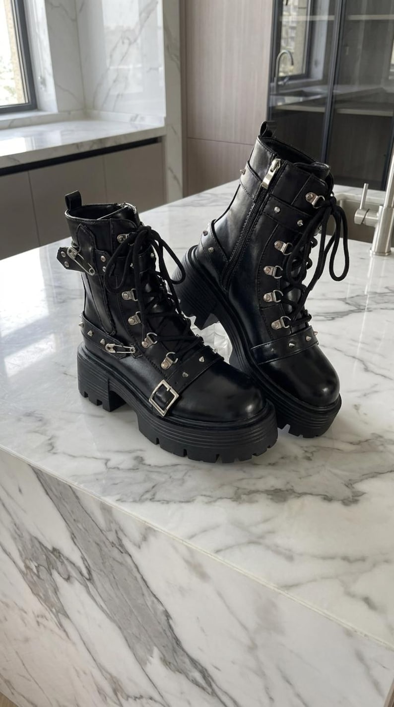 May include: A pair of black leather ankle boots with chunky soles. The boots feature multiple silver-tone metal accents, including buckles, rivets, and eyelets for the laces. The boots are displayed on a marble surface.