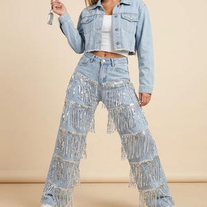 May include: Light blue denim jacket and jeans set. The cropped jacket and high-waisted jeans are adorned with silver fringe. The jeans have multiple rows of fringe cascading down the legs. White sneakers complete the outfit.