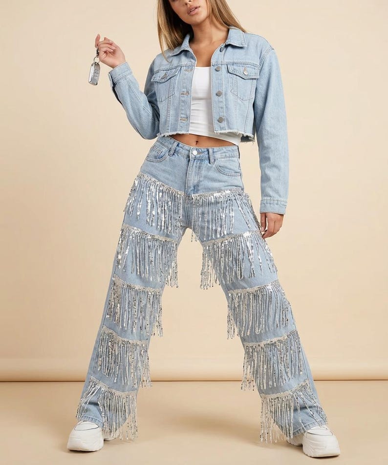 May include: Light blue denim jacket and jeans set. The cropped jacket and high-waisted jeans are adorned with silver fringe. The jeans have multiple rows of fringe cascading down the legs. White sneakers complete the outfit.