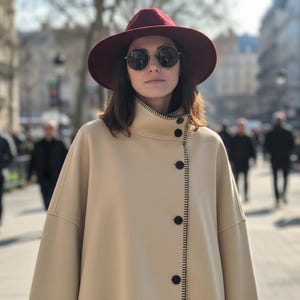 May include: A woman wearing a burgundy fedora hat and round sunglasses. She is wearing a beige coat with black buttons and a high collar. The coat has black stitching along the edges. People are walking in the background.