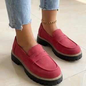 May include: Red loafers with a chunky black sole. The shoes have a classic penny loafer design with a stitched vamp and a rounded toe. The shoes are made of a suede-like material. The shoes are worn with light blue jeans.
