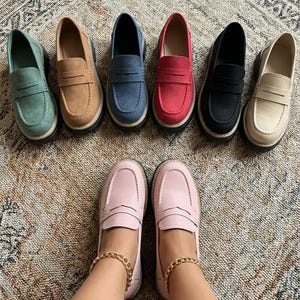 May include: A selection of penny loafers in a range of colours: green, tan, blue, red, black, and beige. The shoes are displayed on a patterned rug, with a pair of pink loafers at the bottom. The shoes have a classic design with a thick sole.