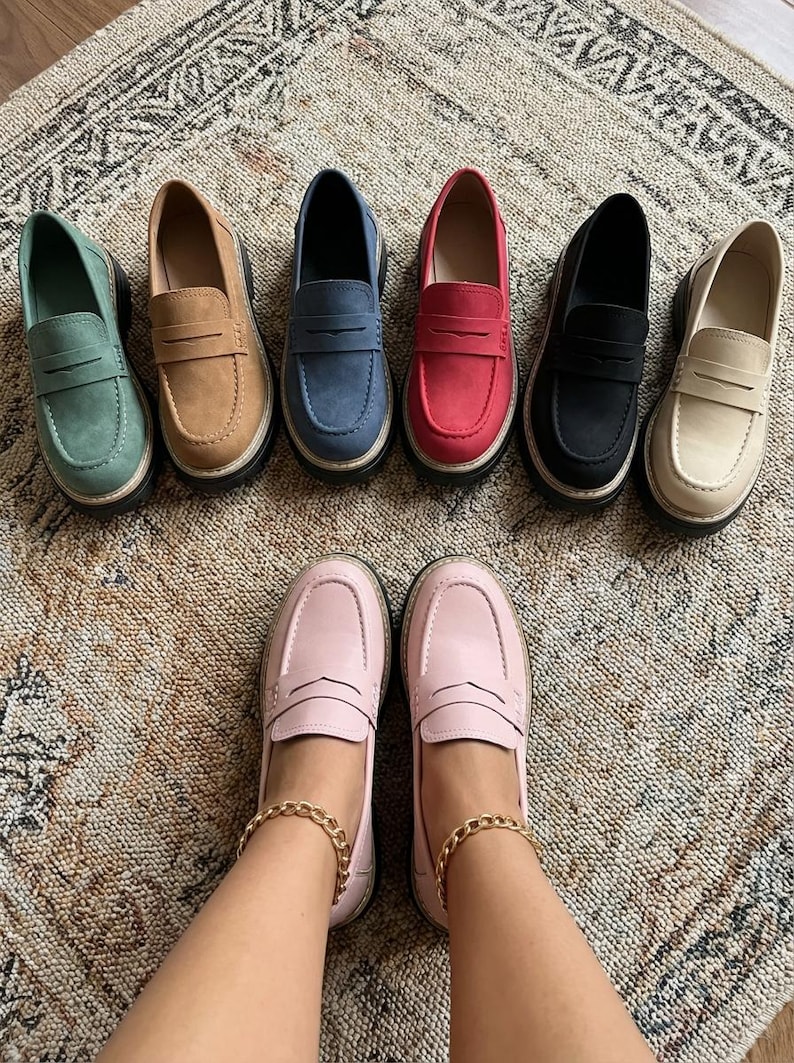 May include: A selection of penny loafers in a range of colours: green, tan, blue, red, black, and beige. The shoes are displayed on a patterned rug, with a pair of pink loafers at the bottom. The shoes have a classic design with a thick sole.