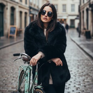 May include: A woman wearing a black faux fur coat and round sunglasses stands next to a vintage teal bicycle on a cobblestone street. The coat has a wide collar and a soft texture. The bicycle has a brown leather seat.