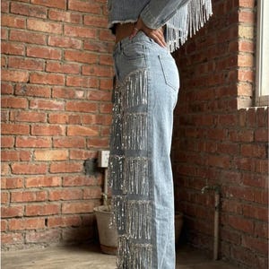 May include: Light blue denim jacket and jeans set with silver fringe detailing. The cropped jacket and wide-leg jeans feature rows of shimmering fringe. The outfit is paired with white platform sneakers, creating a stylish, eye-catching ensemble.