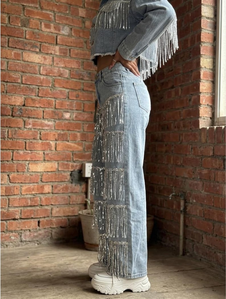 May include: Light blue denim jacket and jeans set with silver fringe detailing. The cropped jacket and wide-leg jeans feature rows of shimmering fringe. The outfit is paired with white platform sneakers, creating a stylish, eye-catching ensemble.