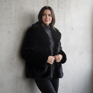 May include: A black faux fur coat with a wide collar and long sleeves. The coat is buttoned and worn over a gray turtleneck. The model is leaning against a concrete wall.