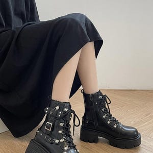 May include: Black combat boots with silver-tone hardware, including buckles, studs, and eyelets. The boots have thick, lug soles and lace-up and zip closures. The boots are worn with a black dress.