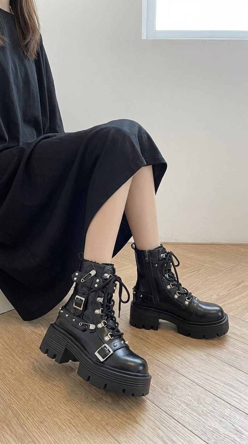 May include: Black combat boots with silver-tone hardware, including buckles, studs, and eyelets. The boots have thick, lug soles and lace-up and zip closures. The boots are worn with a black dress.
