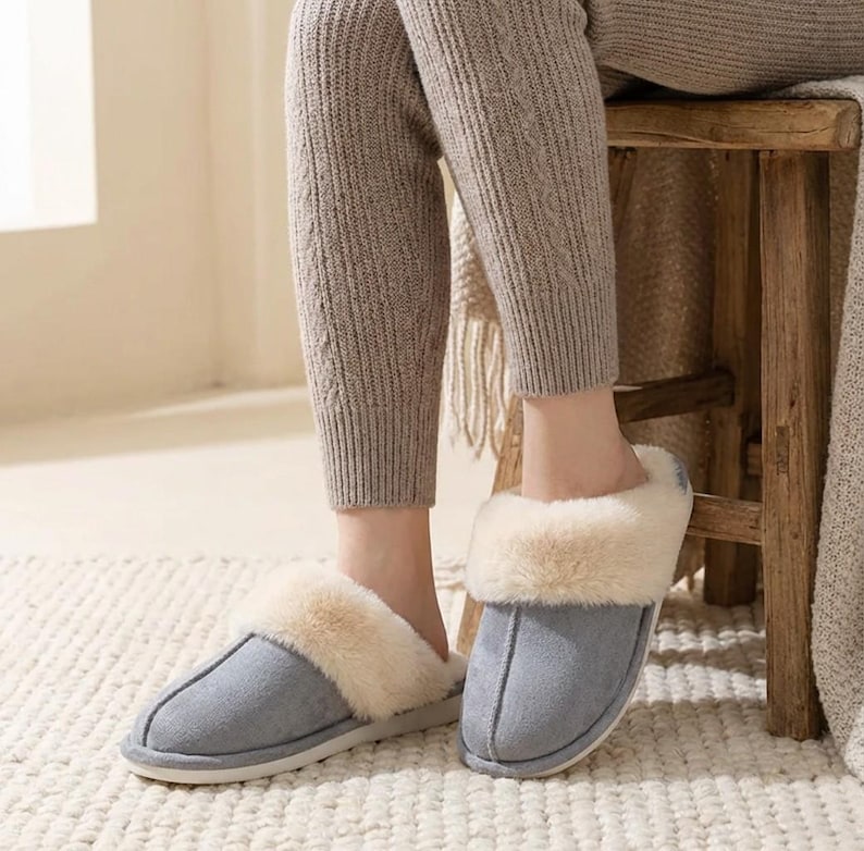Peut inclure: Une paire de pantoufles en daim gris avec une doublure en fausse fourrure cr&egrave;me. Les pantoufles ont un bout arrondi et une semelle blanche. L'image montre les pantoufles port&eacute;es &agrave; l'int&eacute;rieur.