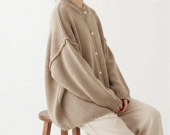 May include: A beige, button-down cardigan sweater with a round neckline. The sweater has a loose fit and is made of a chunky knit material. The sleeves are long, and the buttons are a light beige color. The person is wearing beige pants.