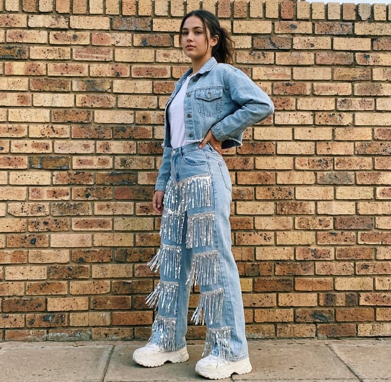 May include: Light blue denim jacket and jeans with silver fringe detailing. The outfit includes a white t-shirt and white sneakers. The background is a brick wall.