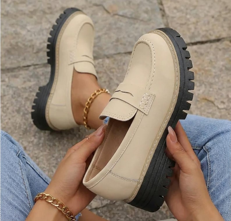 May include: Beige loafers with a black, chunky sole. These shoes feature a classic penny loafer design with a stitched upper and a rounded toe. The soles are approximately 3.8 cm thick. The shoes are shown on a person wearing blue jeans.