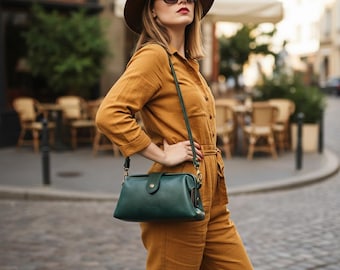May include: A woman wearing a mustard-colored jumpsuit, brown hat, and sunglasses, carrying a dark green leather crossbody bag with a braided strap. The bag has a rectangular shape and a flap closure. The background shows a European street scene.