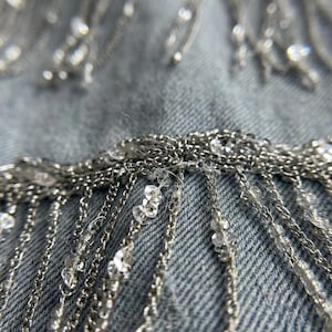 May include: Close-up of a denim fabric embellished with silver chains and clear, sparkling beads. The chains hang in vertical strands, creating a fringe effect. The beads are scattered across the fabric and chains.
