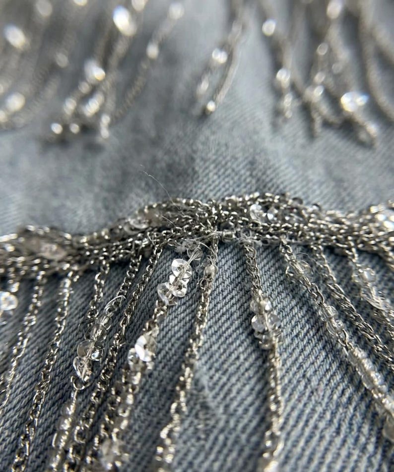 May include: Close-up of a denim fabric embellished with silver chains and clear, sparkling beads. The chains hang in vertical strands, creating a fringe effect. The beads are scattered across the fabric and chains.