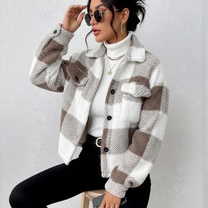 May include: A brown, white, and tan checked jacket with black buttons and two front pockets. The jacket is worn over a white roll-neck. The person is wearing black trousers and sunglasses.