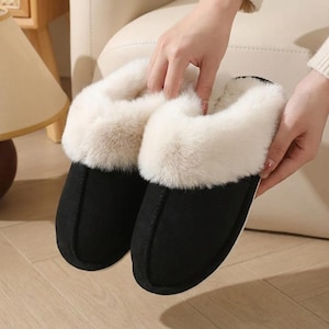 Peut inclure: Une paire de pantoufles noires avec une garniture en fausse fourrure cr&egrave;me. Les pantoufles ont un bout arrondi et une apparence douce et confortable, parfaites pour &ecirc;tre port&eacute;es &agrave; l'int&eacute;rieur. Les pantoufles sont tenues par une personne.