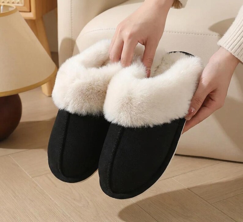Peut inclure: Une paire de pantoufles noires avec une garniture en fausse fourrure cr&egrave;me. Les pantoufles ont un bout arrondi et une apparence douce et confortable, parfaites pour &ecirc;tre port&eacute;es &agrave; l'int&eacute;rieur. Les pantoufles sont tenues par une personne.