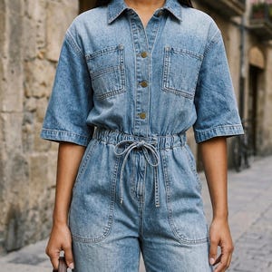 Pode incluir: Um macacão de ganga azul claro com mangas curtas, gola de camisa e fecho frontal de botões. A peça inclui dois bolsos no peito, uma cintura elástica com cordão de ajuste e dois bolsos frontais na zona da anca.