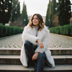 May include: A woman wearing a long, white faux fur coat, gray turtleneck, and dark jeans sits on stone steps. The coat has a wide collar and a long, flowing design. The background features a stone path and tall, green trees.