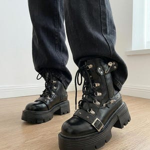 May include: Black combat boots with a chunky platform sole. The boots feature multiple metal eyelets, buckles, and studs. The laces are black, and the boots are worn with black denim jeans.