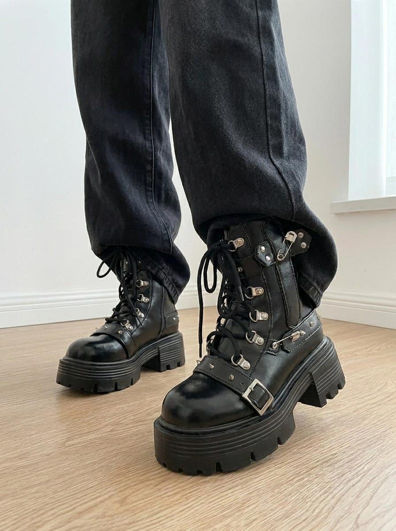 May include: Black combat boots with a chunky platform sole. The boots feature multiple metal eyelets, buckles, and studs. The laces are black, and the boots are worn with black denim jeans.