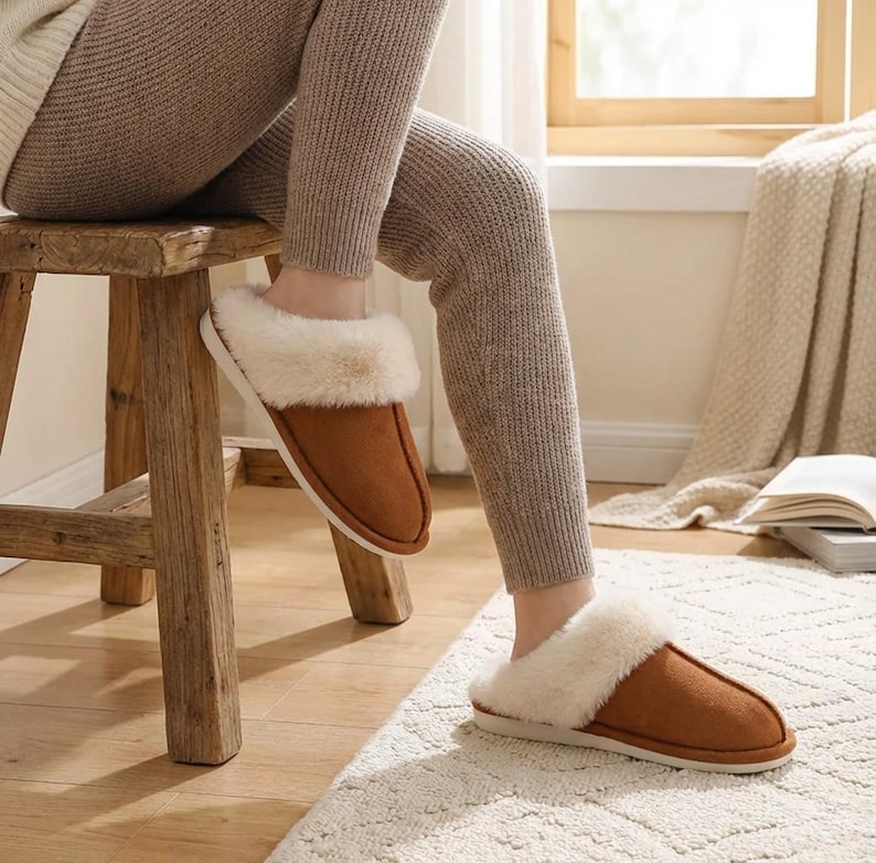 Peut inclure: Pantoufles en daim marron avec une bordure en fausse fourrure cr&egrave;me. Les pantoufles sont pr&eacute;sent&eacute;es sur les pieds d'une personne, pos&eacute;es sur un tapis cr&egrave;me. Les pantoufles ont un aspect doux et confortable.