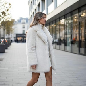 May include: A woman wearing a long, off-white faux fur coat with a wide collar, walking on a city sidewalk. The coat is paired with a white turtleneck and a short skirt. The background features buildings and storefronts.