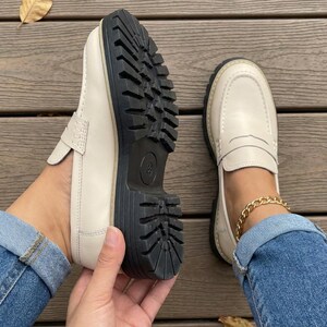 May include: Cream-coloured loafers with a chunky black sole. The shoes have a classic penny loafer design with a stitched upper. The sole is thick and has a tread pattern. The shoes are shown on a wooden surface.
