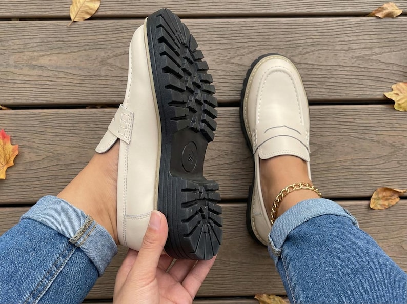 May include: Cream-coloured loafers with a chunky black sole. The shoes have a classic penny loafer design with a stitched upper. The sole is thick and has a tread pattern. The shoes are shown on a wooden surface.