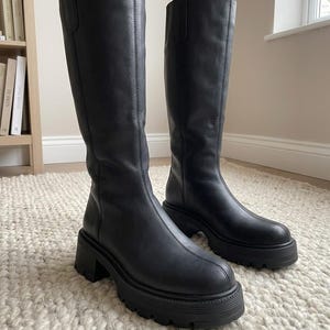 Puede incluir: Un par de botas negras hasta la rodilla con acabado liso, punta redondeada y una suela gruesa con tacos y tacón de bloque. Cada bota tiene una costura vertical en la parte delantera y una trabilla en la parte superior de la caña.