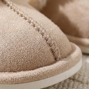 Peut inclure: Gros plan sur une pantoufle beige avec un ext&eacute;rieur textur&eacute; semblable &agrave; du daim. La pantoufle pr&eacute;sente un d&eacute;tail de couture et une semelle blanche. L'accent est mis sur la mati&egrave;re douce et confortable.