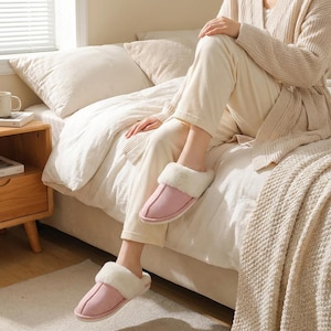Peut inclure: Des chaussons roses avec une bordure en fausse fourrure. Les chaussons sont pos&eacute;s sur un lit avec une literie blanche et une couverture beige. Les chaussons sont con&ccedil;us pour un usage int&eacute;rieur et offrent confort et chaleur.