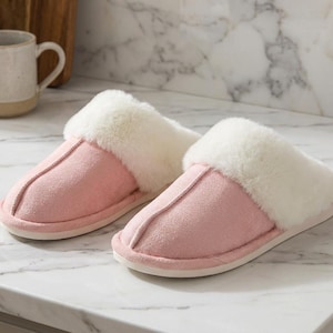 Peut inclure: Une paire de pantoufles en daim rose avec une bordure en fausse fourrure blanche. Les pantoufles sont sur un comptoir en marbre. Une tasse, une planche &agrave; d&eacute;couper et une plante succulente sont en arri&egrave;re-plan.