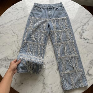 May include: Light blue denim jeans with multiple rows of silver fringe detailing. The jeans have a classic five-pocket design and a straight leg cut. The fringe adds a unique, eye-catching element to the garment.