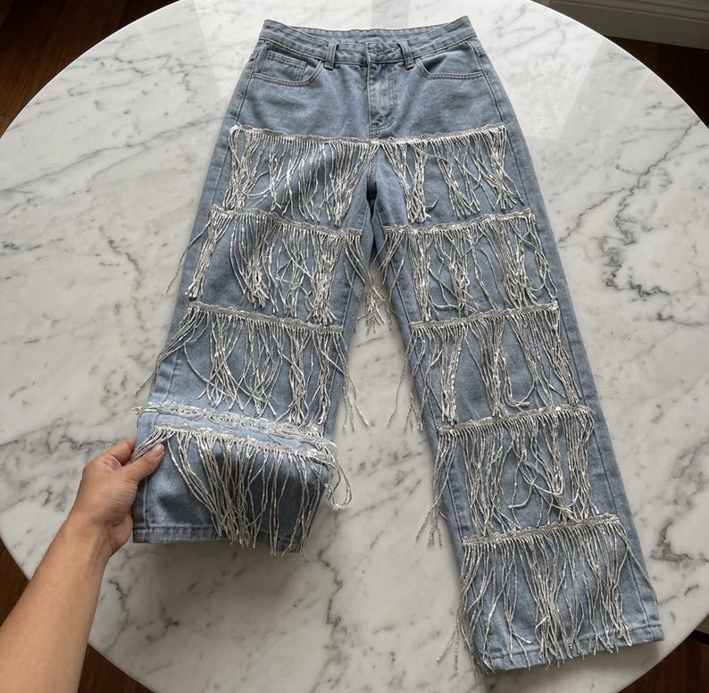 May include: Light blue denim jeans with multiple rows of silver fringe detailing. The jeans have a classic five-pocket design and a straight leg cut. The fringe adds a unique, eye-catching element to the garment.
