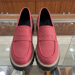 May include: A pair of red loafers with a thick, black and tan sole. The shoes have a classic penny loafer design with a strap across the vamp. The shoes are displayed on a glass surface.