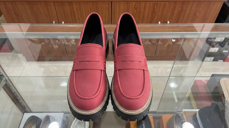 May include: A pair of red loafers with a thick, black and tan sole. The shoes have a classic penny loafer design with a strap across the vamp. The shoes are displayed on a glass surface.
