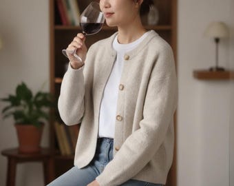 May include: A woman wearing a beige cardigan with wooden buttons, a white t-shirt, and blue jeans. She is holding a glass of red wine. A wooden bookshelf and a small table are in the background.