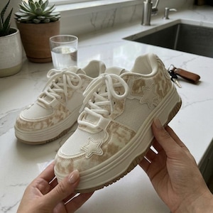 May include: A pair of platform sneakers featuring a white mesh and beige tie-dye patterned upper. The shoes have white laces, a thick white rubber sole, and a prominent star-shaped appliqué on the side and near the toe area.