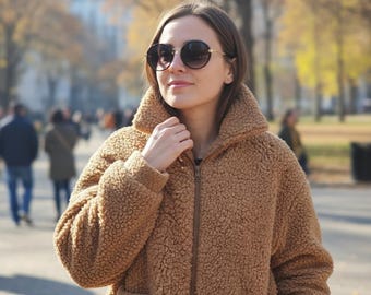 May include: A woman wearing a brown teddy bear coat with a high collar and a zipper closure. She is also wearing dark sunglasses with a gold frame. The background shows a park with trees and people.