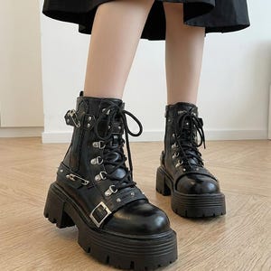 May include: Black platform boots with silver-tone hardware, including buckles, rivets, and eyelets. The boots have a lace-up closure and a chunky, lug sole. The ankle boots are a stylish, edgy footwear choice.