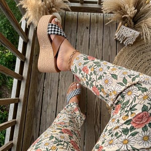 May include: Platform sandals with a black and white houndstooth pattern on the straps and a woven jute sole. The sandals are paired with floral print pants featuring daisies and roses. A gold chain ankle bracelet is also visible.